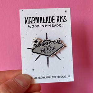 May include: A wooden pin badge with the text "MARMALADE KISS" and "WELCOME to The BLACK PARADE". The pin is shaped like a sign with a starburst design and is held against a white card.