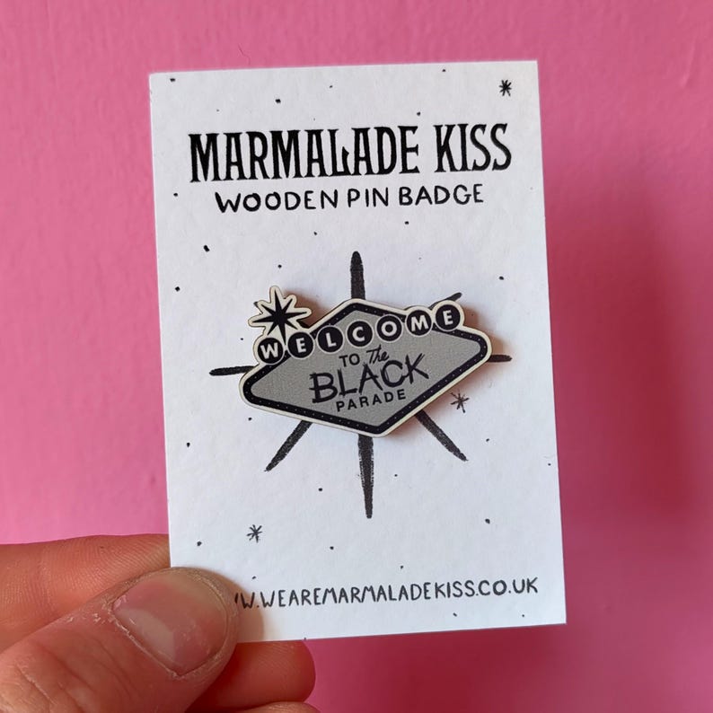 May include: A wooden pin badge with the text "MARMALADE KISS" and "WELCOME to The BLACK PARADE". The pin is shaped like a sign with a starburst design and is held against a white card.