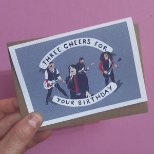 May include: A birthday card featuring a band illustration. The card has a gray background with the text "THREE CHEERS FOR YOUR BIRTHDAY" in a banner. The illustration depicts four band members playing instruments.