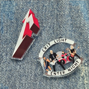 May include: Two acrylic pins. One features a red, white, and brown logo, the other a band illustration with the words "EXIT LIGHT ENTER NIGHT". The band pin shows musicians playing instruments. The pins are on a denim background.