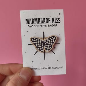 May include: Wooden pin badge featuring a black and white checkered butterfly design. The badge is on a white card with the text "Marmalade Kiss Wooden Pin Badge" and "www.wearemarmaladekiss.co.uk".