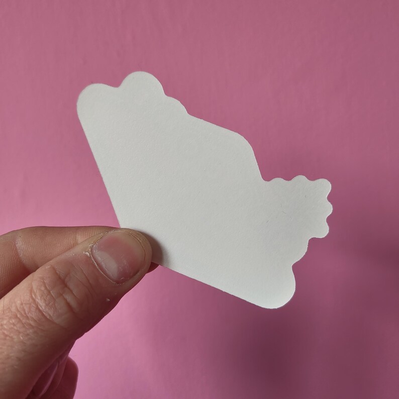 K&ouml;nnte beinhalten: Ein wei&szlig;er Papierauschnitt in Wolkenform wird vor einem rosa Hintergrund gehalten. Der Ausschnitt hat eine glatte, abgerundete Form mit einem gewellten Rand. Das Papier ist schlicht und ohne Verzierungen.