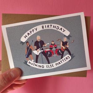 May include: A birthday card featuring a band illustration with the text "HAPPY BIRTHDAY" and "NOTHING ELSE MATTERS". The card has a gray background with a dotted pattern and is held against a pink background.