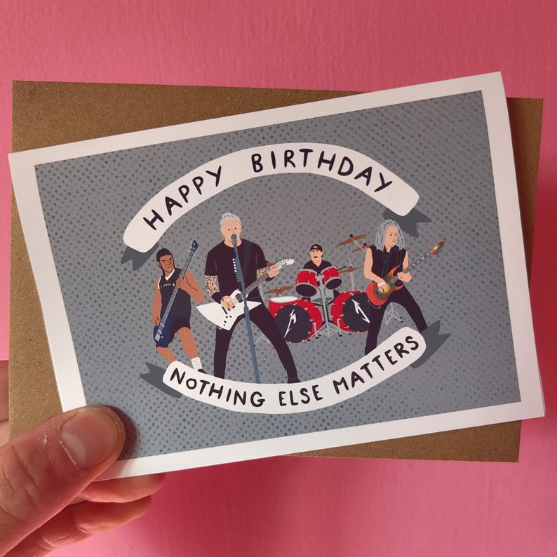 May include: A birthday card featuring a band illustration with the text "HAPPY BIRTHDAY" and "NOTHING ELSE MATTERS". The card has a gray background with a dotted pattern and is held against a pink background.