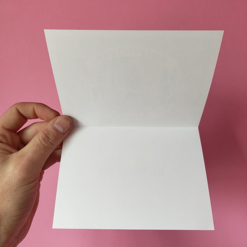 May include: A white greeting card, open to reveal a blank interior, held against a pink background. The card's edges are crisp and clean, with a visible crease down the center. The card is held open by a hand.