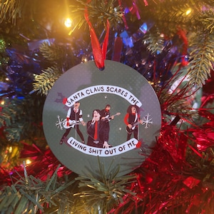 Könnte beinhalten: Ein runder Weihnachtsschmuck mit grünem Hintergrund und dem Text "SANTA CLAUS SCARES THE LIVING SHIT OUT OF ME." Der Schmuck zeigt eine Bandillustration. Ein rotes Band ist zum Aufhängen befestigt.