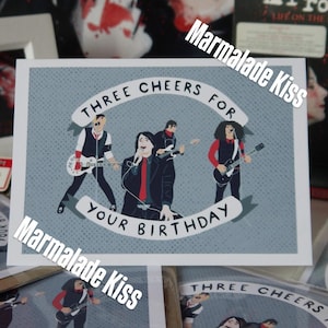 Könnte beinhalten: Eine rechteckige Grußkarte mit hellblauem, gepunktetem Hintergrund und einer stilisierten Illustration einer vierköpfigen Rockband. Der Text "THREE CHEERS FOR YOUR BIRTHDAY" ist in Schwarz auf weißen Bannern über und unter der Band gedruckt.