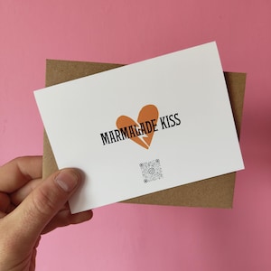 May include: A white greeting card with the text "MARMALADE KISS" and a broken orange heart graphic. The card is held in front of a brown envelope. A QR code is printed below the text. The background is pink.