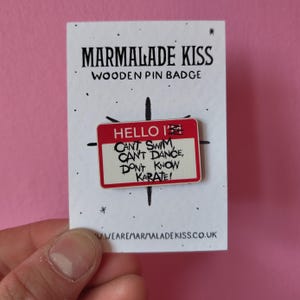 May include: A wooden pin badge with the text "HELLO I CAN'T SWIM, CAN'T DANCE, DON'T KNOW KARATE!" in black and red. The pin is rectangular with a red border and is on a white card with the words "MARMALADE KISS WOODEN PIN BADGE".