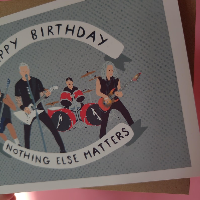 May include: A birthday card featuring a band illustration. The card has a gray background with a dotted pattern. The band members are depicted playing instruments. The text on the card reads "Happy Birthday" and "Nothing Else Matters."