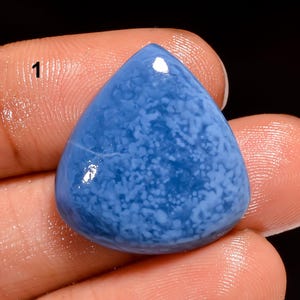 Puede incluir: Una gema pulida en forma de lágrima, de color azul claro, con un patrón de motas blancas. La piedra se sostiene entre dos dedos. El número "1" es visible en la esquina superior izquierda.