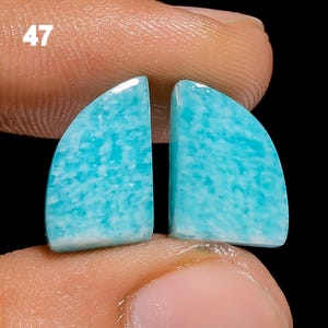 May include: Two light blue and white amazonite cabochons, each shaped like a half-circle. The stones are held in a hand, with the number 47 visible in the background.