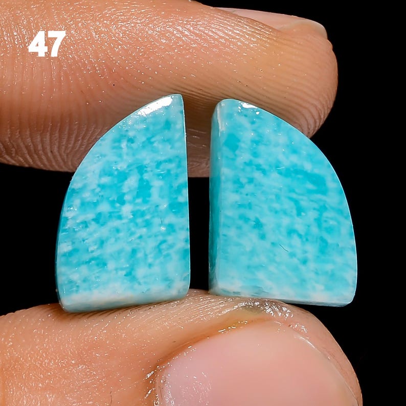 May include: Two light blue and white amazonite cabochons, each shaped like a half-circle. The stones are held in a hand, with the number 47 visible in the background.