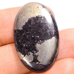 Puede incluir: Una gema de pirita de forma ovalada, con una base oscura casi negra y parches de tonos metálicos dorados y plateados. La piedra tiene una superficie pulida y reflectante, que muestra variaciones naturales de color y textura.