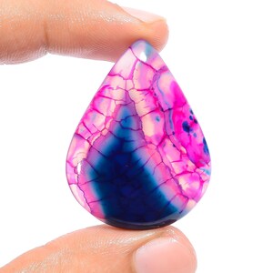 May include: A teardrop-shaped cabochon gemstone with a pink, blue, and white agate pattern. The stone has a smooth, polished surface and is translucent.