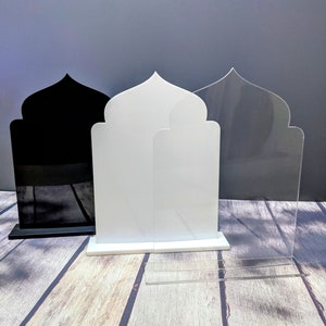 May include: Three arch-shaped display stands in black, white, and clear acrylic. The stands have a distinctive pointed top design and are displayed on a wooden surface. The black and white stands have a solid base, the clear one has a separate base.