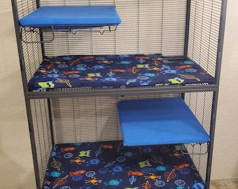 May include: A tall, dark gray metal pet cage with three levels. Each level has a blue cushion or pad. The lower two levels have a dark blue patterned fabric with colorful cartoon space creatures. The cage has wheels.