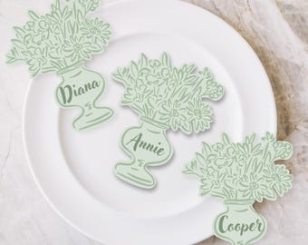 May include: Three light green floral vase place cards with names "Diana", "Annie", and "Cooper" on a white plate. The vases are filled with flowers and have a decorative shape.