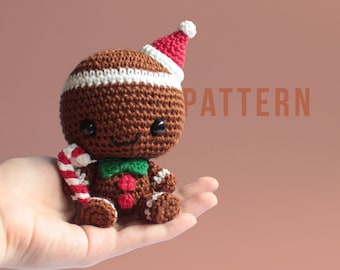 May include: A crocheted gingerbread man toy wearing a red and white Santa hat and holding a candy cane. The toy is sitting on a hand with a brown background. PATTERN is written in the background.