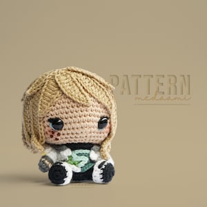 May include: Crocheted amigurumi doll with blonde hair, a green and white outfit, and large black eyes. The doll is sitting on a beige background with the text "PATTERN meddaami" above it.