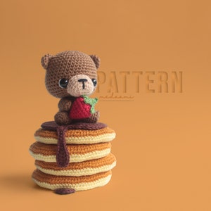 May include: A brown crocheted bear holding a red strawberry sits on top of a stack of four crocheted pancakes with brown chocolate syrup. The text "PATTERN medasami" is in the background.