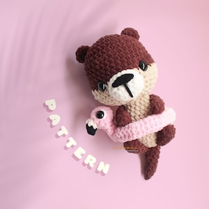 Puede incluir: Una nutria de crochet marrón sosteniendo un flotador de flamenco rosa de crochet. La nutria tiene una cara blanca con ojos negros y una nariz negra. La palabra "PATTERN" está escrita en letras blancas sobre un fondo rosa.