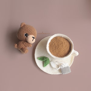 Puede incluir: Un oso de crochet marr&oacute;n sentado junto a una taza de t&eacute; y un platillo de crochet con una bolsita de t&eacute;. La taza de t&eacute; y el platillo son blancos con hojas verdes y una flor blanca.
