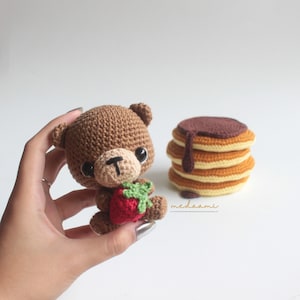 Puede incluir: Un oso de peluche de crochet marr&oacute;n que sostiene una fresa de crochet roja. El oso tiene ojos negros y una nariz negra. El oso est&aacute; sentado en una mano. En el fondo, hay una pila de panqueques de crochet con jarabe de chocolate.