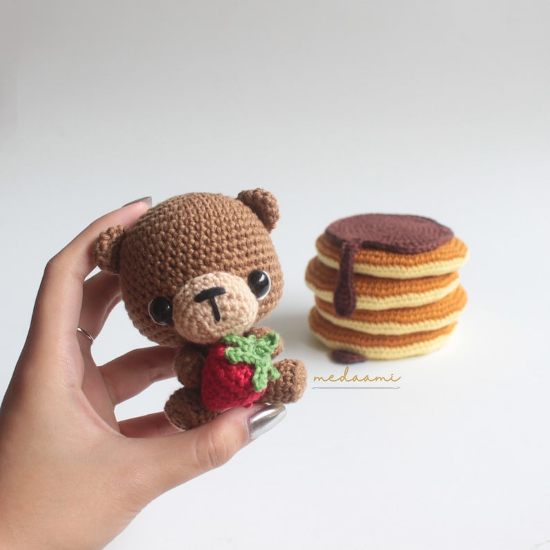 Puede incluir: Un oso de peluche de crochet marr&oacute;n que sostiene una fresa de crochet roja. El oso tiene ojos negros y una nariz negra. El oso est&aacute; sentado en una mano. En el fondo, hay una pila de panqueques de crochet con jarabe de chocolate.