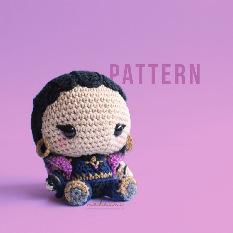 May include: Crochet pattern for a small, brown-skinned amigurumi doll with black hair, wearing a blue and purple outfit. The doll has large, black eyes and a pink blush. The pattern is titled "PATTERN".