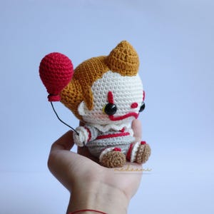 Peut inclure: Un jouet en peluche en crochet repr&eacute;sentant un clown avec des cheveux roux, une chemise ray&eacute;e blanche et rouge et un ballon rouge. Le clown est assis les jambes crois&eacute;es et a une expression triste sur le visage.