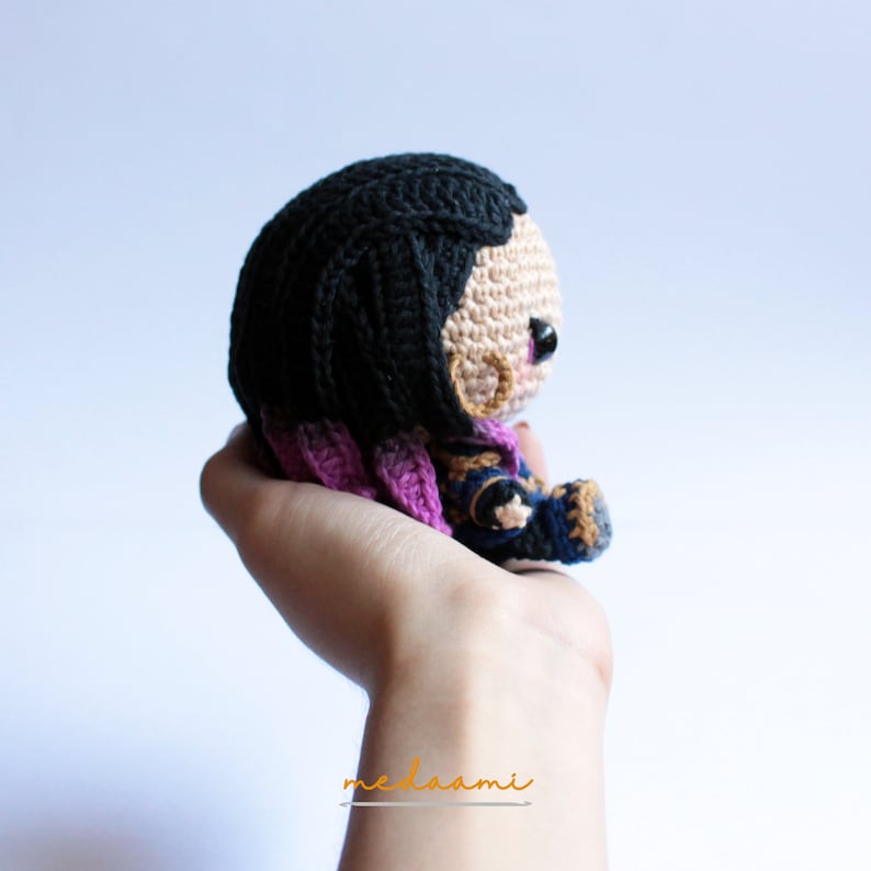 May include: A crocheted doll with black hair, purple and blue clothing, and a gold earring. The doll is sitting in the palm of a hand.