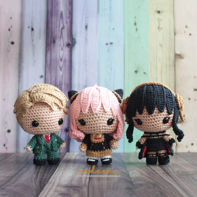 May include: Three crocheted dolls with different hair colors and outfits. The doll on the left has blonde hair and is wearing a green suit. The doll in the middle has pink hair and is wearing a black and gold dress. The doll on the right has black hair and is wearing a black dress with red accents.