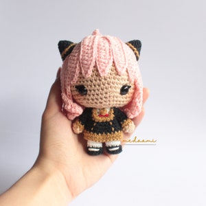 May include: A crocheted doll with pink hair, black cat ears, and a black and gold dress. The doll is holding a small red heart in its hands.