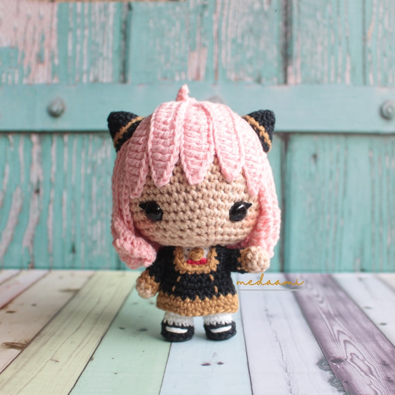 May include: A crocheted amigurumi doll with pink hair, black cat ears, and a black and gold dress. The doll is standing on a wooden surface.