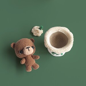 Puede incluir: Un oso de crochet marr&oacute;n con ojos negros y una nariz marr&oacute;n. El oso est&aacute; sentado sobre una superficie verde. Hay una taza de crochet blanca con una hoja verde en la parte superior de la taza. La taza est&aacute; sentada sobre una superficie verde.
