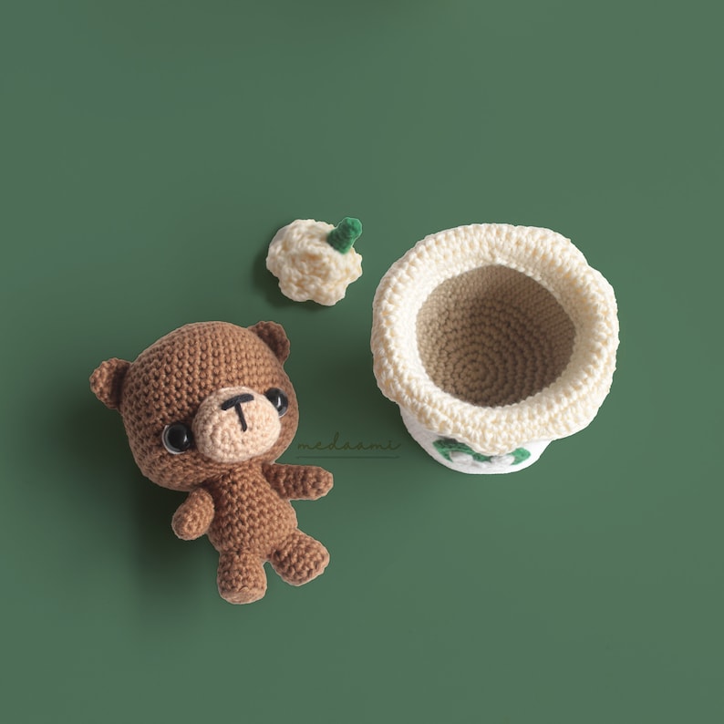 Puede incluir: Un oso de crochet marr&oacute;n con ojos negros y una nariz marr&oacute;n. El oso est&aacute; sentado sobre una superficie verde. Hay una taza de crochet blanca con una hoja verde en la parte superior de la taza. La taza est&aacute; sentada sobre una superficie verde.