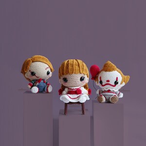 Peut inclure: Trois peluches en crochet, chacune repr&eacute;sentant un personnage de film d'horreur diff&eacute;rent. La premi&egrave;re est un gar&ccedil;on habill&eacute; en rouge et bleu avec un visage cousu, la deuxi&egrave;me est une fille en robe blanche avec des bordures rouges, et la troisi&egrave;me est un clown avec un ballon rouge et une chemise ray&eacute;e.