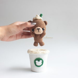 Puede incluir: Un oso de crochet marr&oacute;n que lleva un gorro de crochet blanco con un tallo verde en la parte superior. El oso est&aacute; sosteniendo sus patas en el aire. El oso est&aacute; sentado encima de una taza de caf&eacute; de crochet blanco con un logotipo verde de dos orejas de oso.