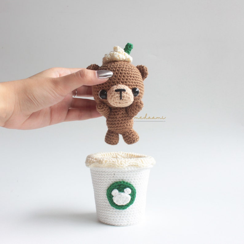 Puede incluir: Un oso de crochet marr&oacute;n que lleva un gorro de crochet blanco con un tallo verde en la parte superior. El oso est&aacute; sosteniendo sus patas en el aire. El oso est&aacute; sentado encima de una taza de caf&eacute; de crochet blanco con un logotipo verde de dos orejas de oso.
