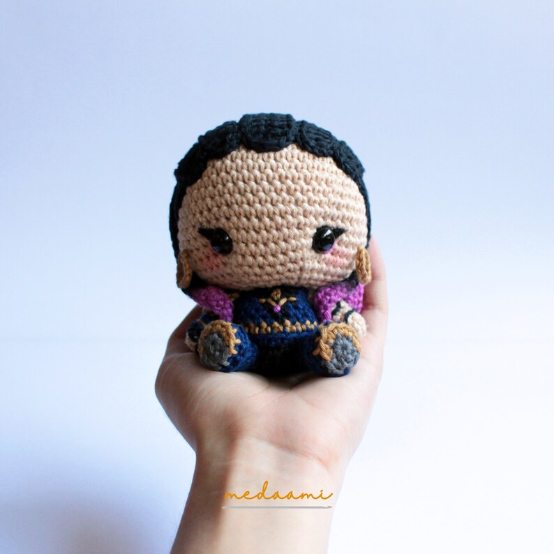 May include: A crocheted amigurumi doll with black hair, a blue and gold outfit, and pink accents. The doll has a sweet expression with large eyes and a small smile.