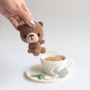 Puede incluir: Un oso de peluche de crochet marr&oacute;n con ojos negros y una nariz marr&oacute;n se sostiene sobre una taza de t&eacute; y un platillo de crochet blanco. La taza de t&eacute; contiene l&iacute;quido marr&oacute;n y una bolsita de t&eacute; de crochet blanco cuelga del borde de la taza.