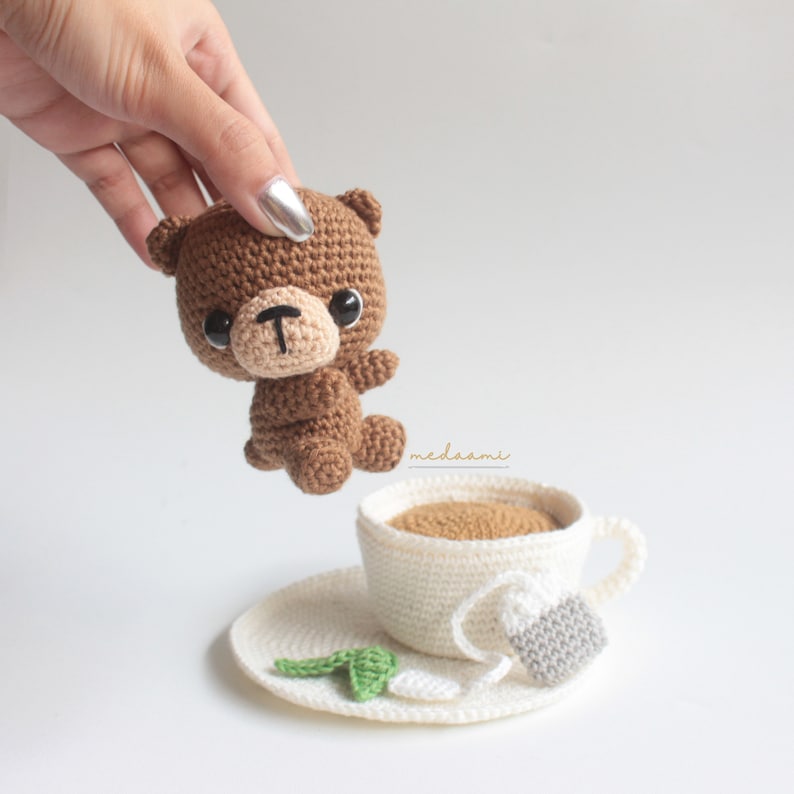 Puede incluir: Un oso de peluche de crochet marr&oacute;n con ojos negros y una nariz marr&oacute;n se sostiene sobre una taza de t&eacute; y un platillo de crochet blanco. La taza de t&eacute; contiene l&iacute;quido marr&oacute;n y una bolsita de t&eacute; de crochet blanco cuelga del borde de la taza.