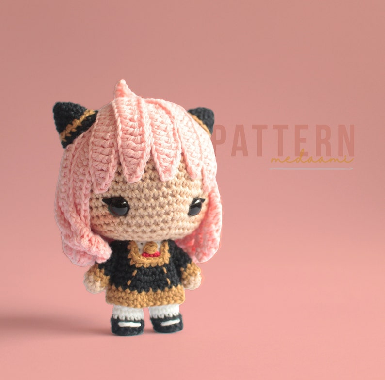 May include: A crocheted amigurumi doll with pink hair, black ears, and a black and brown dress. The doll is standing on a pink background. The text "PATTERN meddaami" is visible in the upper right corner of the image.