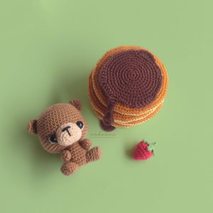 Puede incluir: Un oso de peluche de crochet marr&oacute;n sentado junto a un panqueque de crochet con jarabe de chocolate. Una peque&ntilde;a fresa de crochet roja se encuentra en la esquina inferior derecha de la imagen.