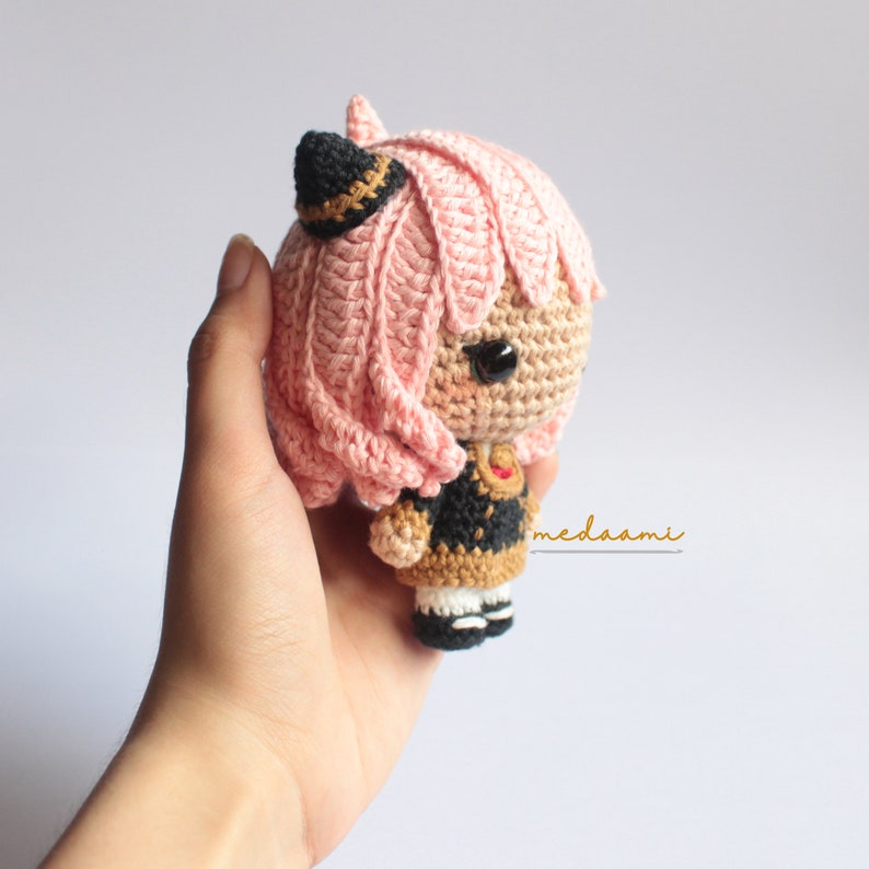 May include: A pink and black crocheted amigurumi doll with a black hat and a gold and black dress. The doll has large black eyes and a pink heart on its chest.