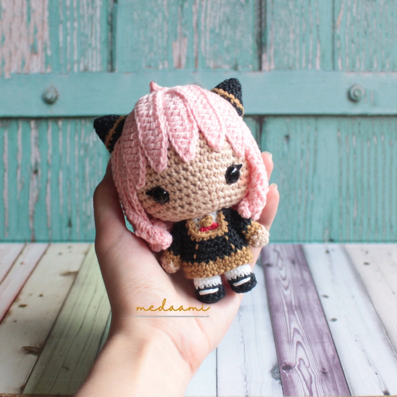 May include: A crocheted doll with pink hair, black cat ears, and a black and gold dress. The doll is holding a small, black and gold object in its hands.