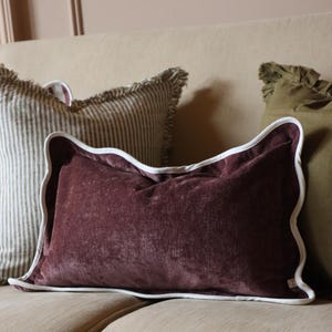 May include: A rectangular plum-coloured velvet cushion with a white scalloped edge. The cushion is the main focus, resting on a beige sofa. Other cushions with striped and ruffled textures are also visible in the image.