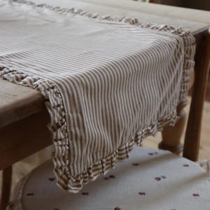 May include: A beige and brown striped table runner with ruffled edges. The runner is draped over a wooden table, showcasing its design and texture. The fabric has a classic, farmhouse aesthetic.