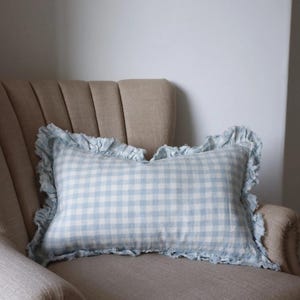 Könnte beinhalten: Ein rechteckiges Zierkissen mit hellblauem und weißem Vichy-Muster. Das Kissen hat einen gerüschten Rand und liegt auf einem beige gepolsterten Sessel. Das Kissen verleiht dem Raum einen Hauch von Landhausstil.